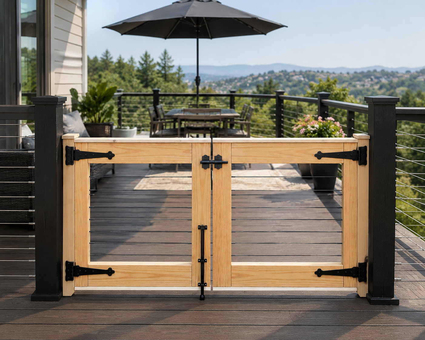 A light natural wood deck gate with black metal hinges and latch hardware, installed on a dark wood deck with a patio umbrella and outdoor landscape in the background.